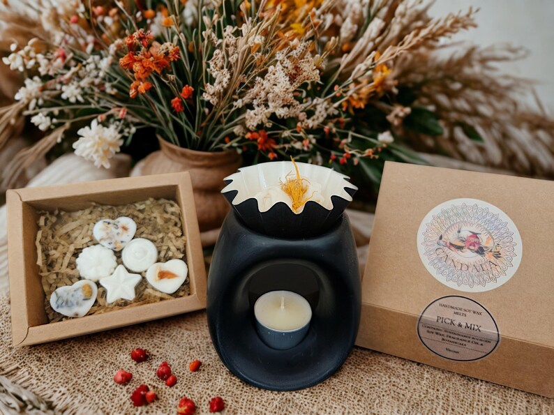 May include: A black essential oil wax melt warmer with a lit tealight candle inside. A small wooden box contains wax melts in various shapes. A brown box with a circular sticker is next to the warmer. Dried flowers are in the background.