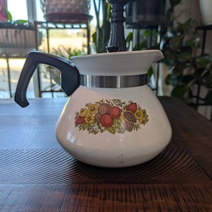 May include: White vintage coffee pot with a black handle and a silver band. The pot features a colorful floral and fruit design. The pot is sitting on a dark wooden surface.