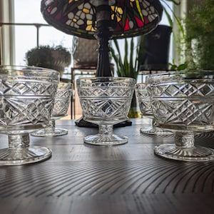 May include: A set of six clear glass dessert dishes with a diamond pattern. The dishes have a pedestal base and a wide, shallow bowl. The glass is clear, and the dishes are arranged on a dark wooden surface.