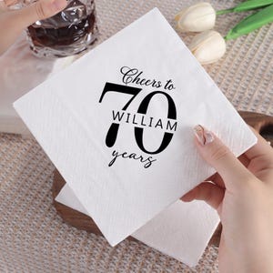 May include: A square white paper napkin featuring black printed text that reads Cheers to 70 WILLIAM years. The napkin has a textured, embossed border pattern along the edges and is held up to display the centred graphic design clearly.