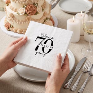 Pode incluir: Um guardanapo de papel branco quadrado com texto impresso em preto que diz Cheers to 70 years WILLIAM. O guardanapo tem um padrão de borda texturizado e em relevo, sendo segurado para exibir a mensagem comemorativa de aniversário.