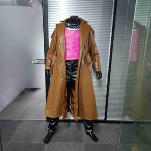 Puede incluir: Un abrigo largo de cuero marrón con una camisa rosa debajo. El abrigo tiene un cinturón y un cuello. La camisa tiene un diseño muscular.
