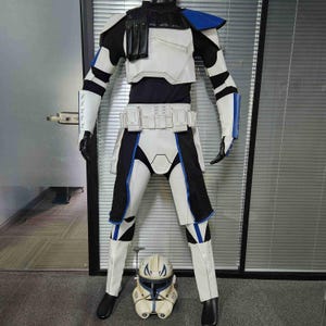 Op de afbeelding: Een volledig clone trooper-kostuum in wit, zwart en blauw. De outfit bevat een borstplaat, riem en beenkappen. Een bijpassende helm staat aan de voeten. Het kostuum is geïnspireerd op een sciencefictionfranchise.