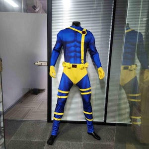 Puede incluir: Un traje de spandex azul y amarillo con un cinturón y correas amarillas. El traje tiene un diseño muscular y un símbolo rojo en el pecho.