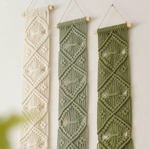 Op de afbeelding: Drie macramé wandkleden in crème, saliegroen en olijfgroen. Elk rechthoekig stuk heeft een geometrisch ruitpatroon en is opgehangen aan een houten deuvel met een koord van natuurlijke vezels. De wanddecoratie voegt een bohemien tintje toe.