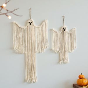 Puede incluir: Dos colgantes de pared de fantasmas de macramé con ojos de cuentas negras. Los fantasmas están hechos de hilo color crema y cuelgan de una pared blanca. El fantasma más grande mide aproximadamente 76 cm de alto.