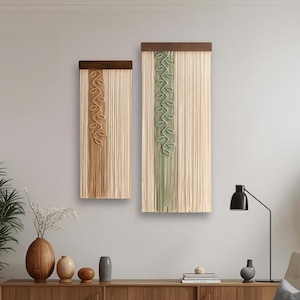 May include: Two macrame wall hangings with wooden tops. One features a tan and brown design, the other a green design. The hangings are displayed on a light grey wall above a wooden cabinet, with plants and a chair.