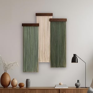 May include: Three wall hangings with a modern design. Each piece features a dark wood top with vertical strands in green, cream, and olive green. The room also includes a wooden cabinet, a chair, and plants, creating a cozy atmosphere.