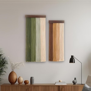 May include: Two rectangular wall hangings with a wooden top and vertical strands in shades of green, beige, and brown. The decor is displayed on a white wall above a wooden cabinet. The room is decorated with plants, vases, and a modern chair.