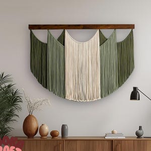 May include: A large macrame wall hanging with a wooden bar, featuring olive green and cream-coloured fringe. The wall decor is mounted on a white wall above a wooden cabinet. The overall theme is bohemian home decor.