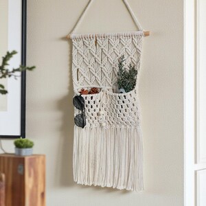 May include: Cream-colored macrame wall hanging with two pockets and a fringe. The top is a triangle shape, and the pockets hold sunglasses, greenery, and decorative items. The wall hanging is a bohemian style.