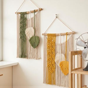 Op de afbeelding: Twee macramé wandkleden met bladvormgeving. Eén is olijfgroen en crème, de andere is mosterdgeel en crème. Elk heeft een houten deuvel en een ophangkoord. De achtergrond is een neutrale muur.