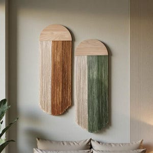 Pode incluir: Duas decorações de parede com tampo de madeira e fundo com franjas. Uma é marrom e bege, a outra é verde e bege. As decorações estão acima de uma cama com roupa de cama bege. Uma lâmpada branca está em uma mesa de cabeceira.