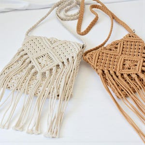 Puede incluir: Dos bolsos cruzados de macramé en crema y marrón claro. Los bolsos presentan intrincados patrones tejidos y largos flecos. Cada bolso tiene una correa trenzada para llevar al hombro. Los bolsos se muestran sobre una superficie blanca.