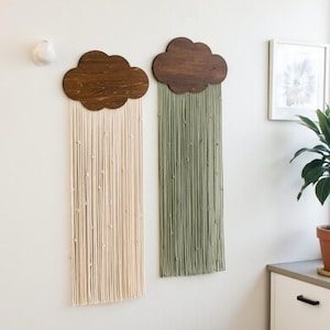 Puede incluir: Dos colgantes de pared con forma de nube. Las nubes son de madera oscura, con hilo de color crema y verde oliva colgando por debajo. Los colgantes de pared están montados en una pared blanca. También se ve un cuadro enmarcado y una planta en maceta.