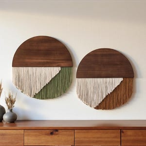 May include: Two circular wall hangings with a dark wood top and a textured, fringed bottom. One features off-white and olive green, the other off-white and brown. The wall art is displayed above a wooden cabinet.