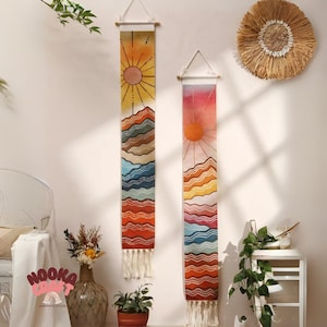 Könnte beinhalten: Zwei vertikale Wandbehänge mit Landschaftsdesign. Jedes zeigt eine Sonne, Berge und farbige Streifen in Orange-, Gelb-, Blau- und Rottönen. Sie bestehen aus Stoff und haben oben Holzstäbe zum Aufhängen, mit Quasten unten.