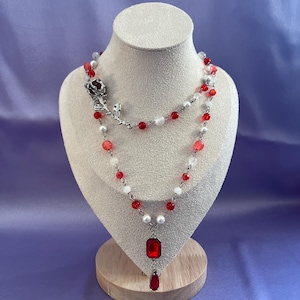 Pode incluir: Um colar em camadas com contas vermelhas e brancas, pérolas e um detalhe de rosa prateada. O colar apresenta um grande pingente vermelho retangular e um pingente vermelho em forma de gota. A joia é exibida em um manequim.