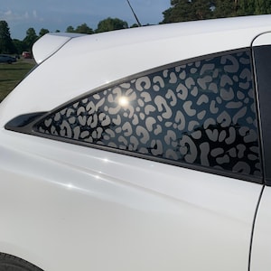 May include: A white car features a rear side window decorated with a dark grey leopard print decal. The pattern consists of various irregular, rounded shapes that cover the glass surface, contrasting against the white body of the vehicle.