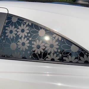 May include: A white car features a triangular rear side window decorated with a repeating vinyl decal pattern of light grey daisies and small circular dots. The floral design covers the glass surface, creating a decorative graphic overlay.