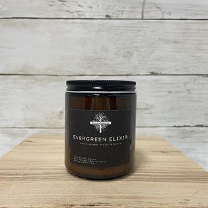 May include: A brown glass jar candle with a black lid and a black label. The label reads "EVERGREEN ELIXIR" with the scent description "alpine balsam, dry gin & cypress". The candle is on a wooden surface.
