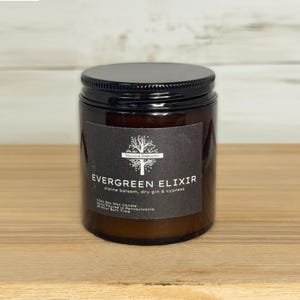 May include: A brown glass jar candle with a black lid. The label reads "Evergreen Elixir" with the scent description. The candle is on a wooden surface.