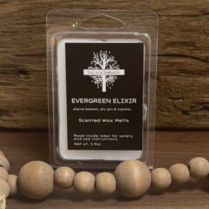 May include: A package of scented wax melts labeled "Evergreen Elixir" with a black and white label. The label includes the text "Sylvan & Simplicity" and lists ingredients. The net weight is 2.9oz. The package is in a clear plastic container.