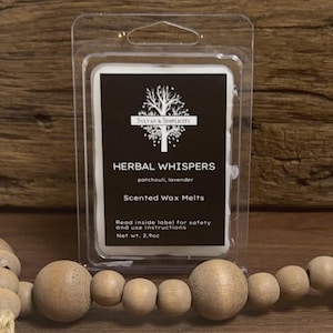 May include: A package of scented wax melts labeled "Herbal Whispers" with patchouli and lavender scents. The black label features a tree design and the text "Sylvan & Simplicity." The net weight is 2.9 oz. The package is in a clear plastic container.