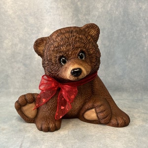 May include: A brown ceramic teddy bear figurine with a red ribbon tied around its neck. The ribbon features small red hearts. The bear is seated with its legs extended and has a sweet expression.