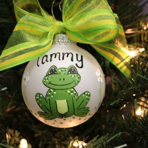 May include: A white Christmas ornament featuring a hand-painted green frog with black eyes and the name "Tammy". The ornament is decorated with small white flowers and a green and yellow ribbon.