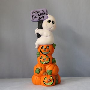 Könnte beinhalten: Eine Halloween-Figur mit einem Stapel orangefarbener Kürbisse mit lächelnden Gesichtern. Eine weiße Geisterfigur sitzt oben auf den Kürbissen und hält ein lila Schild mit der Aufschrift "Have a Boo-tiful Day" in Schwarz.
