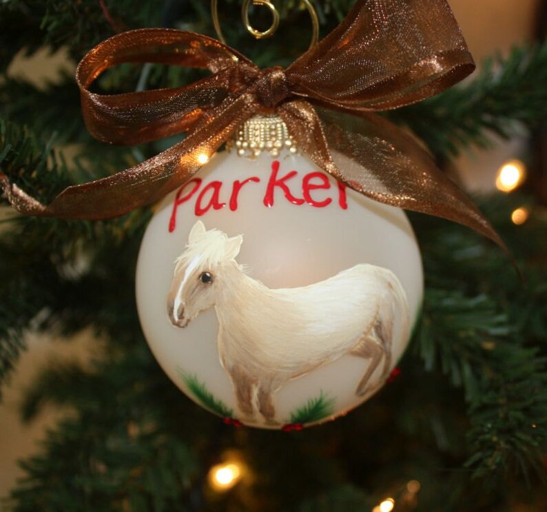 K&ouml;nnte beinhalten: Weihnachtskugel mit handgemaltem Pferd und dem Namen "Parker" in Rot. Verziert mit einem braunen Band, h&auml;ngt sie an einem Weihnachtsbaum und schafft eine festliche Atmosph&auml;re.