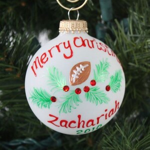 K&ouml;nnte beinhalten: Wei&szlig;er Weihnachtsschmuck mit rotem Text "Merry Christmas" und "Zachariah 2010." Der Schmuck zeigt einen braunen Football, gr&uuml;ne Zweige und rote Schmuckakzente. Oben befindet sich eine goldfarbene Metallschlaufe zum Aufh&auml;ngen.