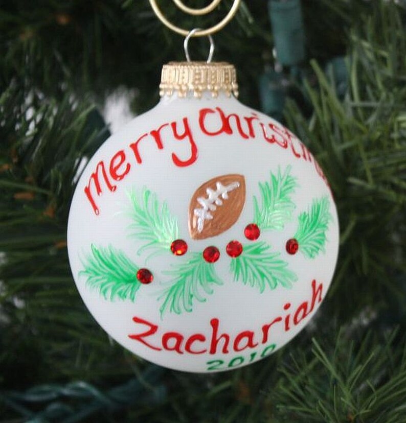 K&ouml;nnte beinhalten: Wei&szlig;er Weihnachtsschmuck mit rotem Text "Merry Christmas" und "Zachariah 2010." Der Schmuck zeigt einen braunen Football, gr&uuml;ne Zweige und rote Schmuckakzente. Oben befindet sich eine goldfarbene Metallschlaufe zum Aufh&auml;ngen.