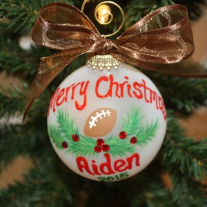 K&ouml;nnte beinhalten: Ein wei&szlig;er Weihnachtsschmuck mit einem Football-Design, den Worten "Merry Christmas" und dem Namen "Aiden" in Rot. Der Schmuck ist mit gr&uuml;nen Tannennadeln und roten Beeren verziert und mit einem braunen Band versehen.