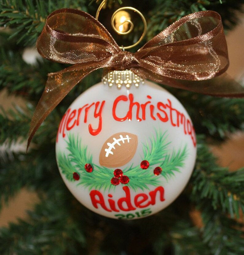 K&ouml;nnte beinhalten: Ein wei&szlig;er Weihnachtsschmuck mit einem Football-Design, den Worten "Merry Christmas" und dem Namen "Aiden" in Rot. Der Schmuck ist mit gr&uuml;nen Tannennadeln und roten Beeren verziert und mit einem braunen Band versehen.