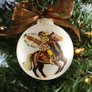 K&ouml;nnte beinhalten: Wei&szlig;er Christbaumschmuck mit einem Motiv eines Indianers auf einem Pferd und den Buchstaben "FSU". Verziert mit einem braunen Band, h&auml;ngt er an einem Weihnachtsbaum. Eine festliche Dekoration.