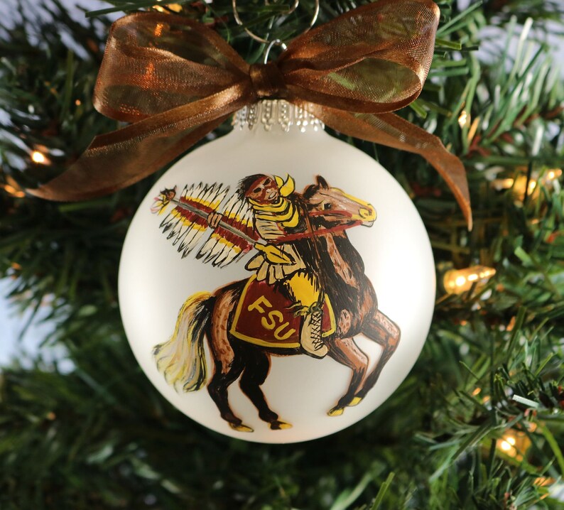 K&ouml;nnte beinhalten: Wei&szlig;er Christbaumschmuck mit einem Motiv eines Indianers auf einem Pferd und den Buchstaben "FSU". Verziert mit einem braunen Band, h&auml;ngt er an einem Weihnachtsbaum. Eine festliche Dekoration.