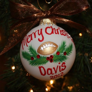 K&ouml;nnte beinhalten: Wei&szlig;er Weihnachtsschmuck mit einem goldenen Football-Design, rotem Text mit der Aufschrift "Merry Christmas" und "Davis 2012". Der Schmuck ist mit gr&uuml;nen Tannennadeln und roten Juwelen verziert und mit einem braunen Band versehen.