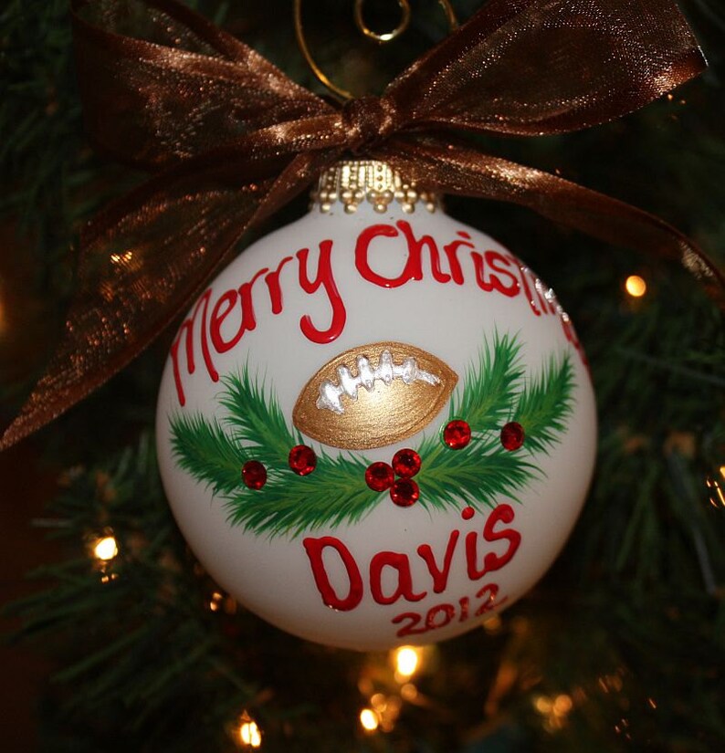K&ouml;nnte beinhalten: Wei&szlig;er Weihnachtsschmuck mit einem goldenen Football-Design, rotem Text mit der Aufschrift "Merry Christmas" und "Davis 2012". Der Schmuck ist mit gr&uuml;nen Tannennadeln und roten Juwelen verziert und mit einem braunen Band versehen.