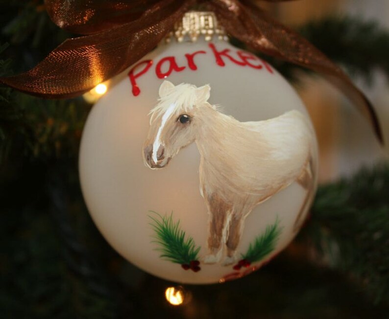 K&ouml;nnte beinhalten: Eine Christbaumkugel aus mattiertem Glas mit einem handgemalten Pferdemotiv. Das Pferd ist hellbraun mit wei&szlig;er M&auml;hne und Schweif. Der Name "Parker" steht in roter Schrift. Oben ist ein braunes Band gebunden.