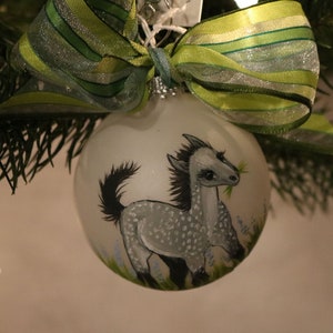 K&ouml;nnte beinhalten: Eine transparente Weihnachtskugel aus Glas mit einem handgemalten grauen Pferdemotiv. Das Pferd hat eine schwarze M&auml;hne und einen schwarzen Schweif und frisst Gras. Die Kugel ist mit einer gr&uuml;n-silbernen Schleife verziert.