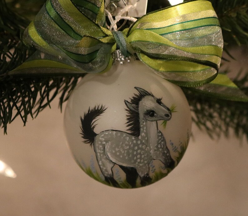 K&ouml;nnte beinhalten: Eine transparente Weihnachtskugel aus Glas mit einem handgemalten grauen Pferdemotiv. Das Pferd hat eine schwarze M&auml;hne und einen schwarzen Schweif und frisst Gras. Die Kugel ist mit einer gr&uuml;n-silbernen Schleife verziert.
