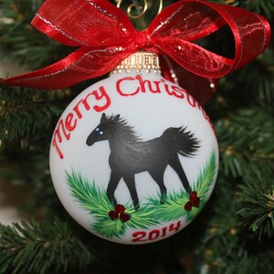 K&ouml;nnte beinhalten: Wei&szlig;e Weihnachtskugel mit einer Illustration eines schwarzen Pferdes, gr&uuml;nem Laub und roten Beeren. Der Schmuck hat die Worte "Merry Christmas" und das Jahr "2014" in Rot. Oben ist ein rotes Band mit silbernem Rand gebunden.