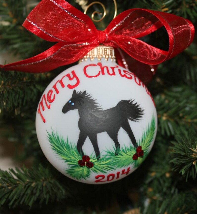 K&ouml;nnte beinhalten: Wei&szlig;e Weihnachtskugel mit einer Illustration eines schwarzen Pferdes, gr&uuml;nem Laub und roten Beeren. Der Schmuck hat die Worte "Merry Christmas" und das Jahr "2014" in Rot. Oben ist ein rotes Band mit silbernem Rand gebunden.