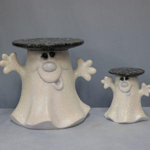 Puede incluir: Dos soportes de cerámica con forma de fantasma y acabado blanco brillante, cada uno con una parte superior redonda de color negro moteado. Las figuras tienen los brazos levantados, ojos pintados y una nariz pequeña. El fantasma grande está a la izquierda.
