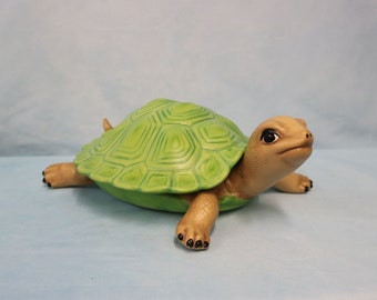 May include: A ceramic turtle figurine with a light green shell and tan head and limbs. The shell has a textured, geometric pattern. The turtle's eyes are black, and its feet have black claws. The figurine is on a light blue surface.
