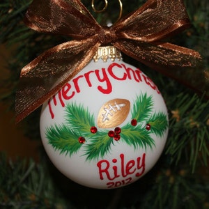 K&ouml;nnte beinhalten: Wei&szlig;er Weihnachtsschmuck mit einem goldenen Football-Design, gr&uuml;nen Tannennadeln und roten Beeren. Der Schmuck tr&auml;gt die Worte "Merry Christmas" und "Riley 2012" in Rot. Eine braune Schleife aus Band ziert die Oberseite.