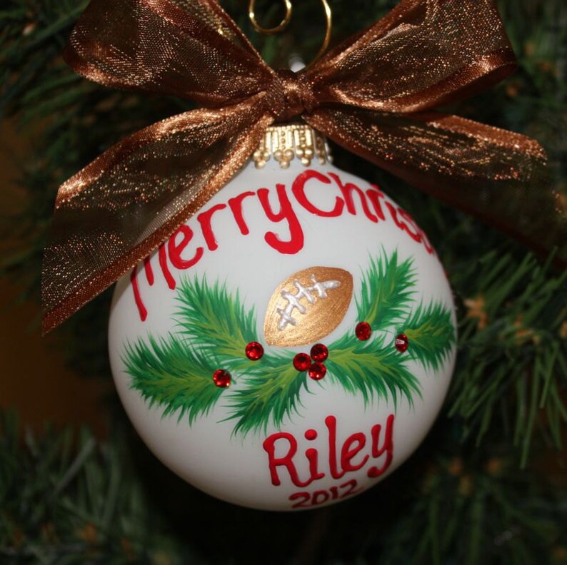 K&ouml;nnte beinhalten: Wei&szlig;er Weihnachtsschmuck mit einem goldenen Football-Design, gr&uuml;nen Tannennadeln und roten Beeren. Der Schmuck tr&auml;gt die Worte "Merry Christmas" und "Riley 2012" in Rot. Eine braune Schleife aus Band ziert die Oberseite.
