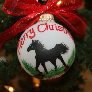 K&ouml;nnte beinhalten: Wei&szlig;er Weihnachtskugel mit einer schwarzen Pferdeillustration und dem Schriftzug "Merry Christmas" in Rot. Der Schmuck hat ein rotes Band und h&auml;ngt an einem Weihnachtsbaum.